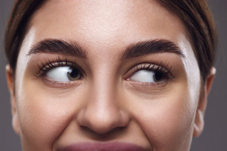 Cropped photo. Close-up of womans eyes with well-groomed eyebrows, clear eyes, and smooth skin against a grey background. Concept of medical, exercises techniques for vision, and focusing for eyes.の写真素材