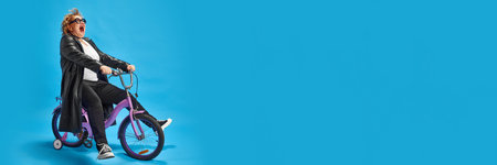 Elderly woman exuding coolness and joy on purple bicycle dressed leather jacket, sunglasses against blue background. Negative space. Concept of fashionable older adults, active seniors in modern life.の写真素材