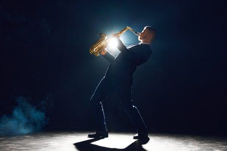 Elegant man with sax tilted upwards performing new melody on stage against dark background with theatrical smoke. Concept of art, instrumental music, dance, culture, festivals and concerts.の写真素材