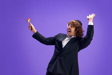 Joyous Jewish man in black suit and quirky glasses holding colorful toy gregger against purple studio background. Celebration Purim, business, festival, holiday, judaism, religion concept. Adの写真素材