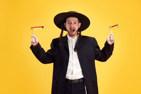 Overjoyed young Jewish man with widely open mouth holds groggers in hands against sunny yellow background. Purim, business, festival, holiday, celebration Pesach or Passover, judaism, religion conceptの写真素材