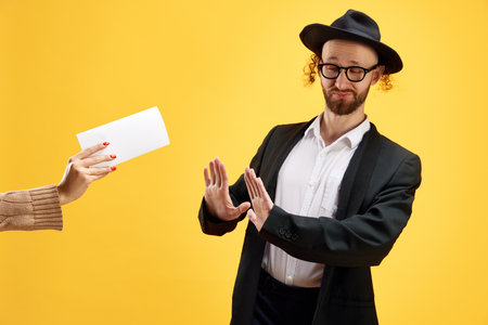 Jewish man refuses envelope with money held by someones hand against warm yellow studio background. Purim, business, festival, holiday, celebration Pesach or Passover, judaism, religion concept.の写真素材