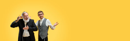 Banner. Two positive young and senior men posing in formal suits against yellow background with negative space to insert text. Purim, business, festival, holiday, celebration concept. Adの写真素材