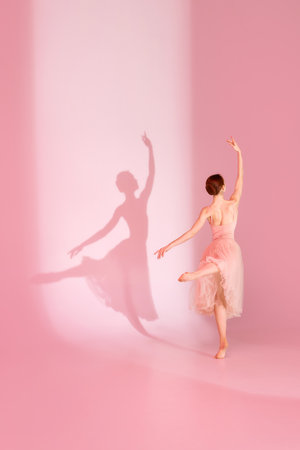 Balletic Beauty. Classic ballerina dances barefoot in pink dress against pastel pink background with her shadow. Concept of art and hobby, elegance, ballet, beauty and femininity.の写真素材