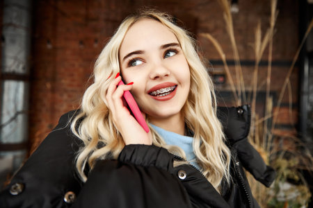 Young blonde woman with dental braces in winter coat stands on terrace and smiling talking on phone. Concept of youth and healthcare, medicine and beauty, confidence and selfcare.の写真素材