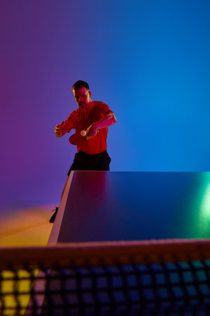 Young man, table tennis competitor in mid-swing during competitive match in neon light against...