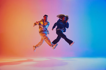 Two dancers in mid-air jump while dancing in motion against gradient blue-orange background in neon lighting. Movement. Concept of youth culture, music, lifestyle, fashion, action. Gel portrait.の写真素材