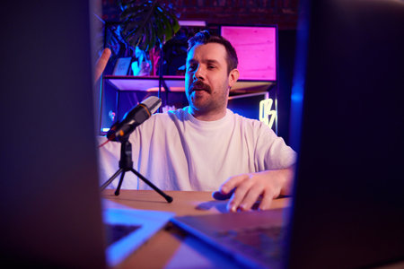 Excited man with microphone in neon-lit home studio engaging with computer screen and creating...