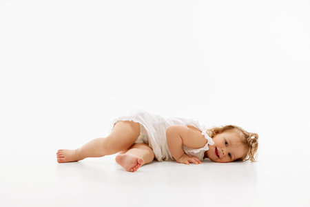 Playful and happy little baby-girl with curly hair lying on floor and laughing in cute white dress against white background. Concept of childhood, motherhood, life, birth. Copy space for adの写真素材