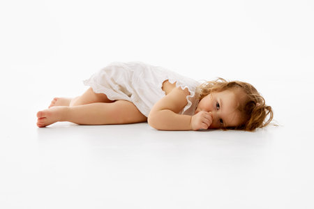 Cute little baby with curly and wet hair in frilly dress resting on floor with thoughtful expression against white background. Concept of childhood, carefree, motherhood, life, birth. Copy space. Adの写真素材