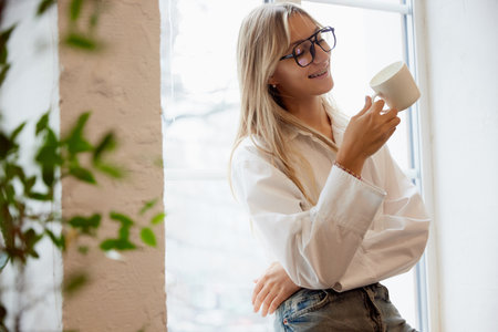 Young woman in eyeglasses drinking coffee by window. Model with perfect smile with braces. Orthodontic Treatment. Dental care. Concept of beauty and medicine, malocclusion, orthodontic health.の写真素材