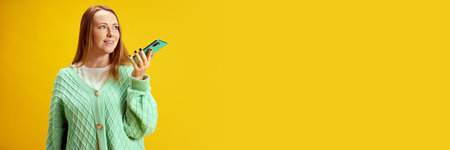 Banner. Young woman using smartphone and speaking by cellular connection against yellow studio background with negative space against studio background. Concept of online working, delivery, technologyの写真素材