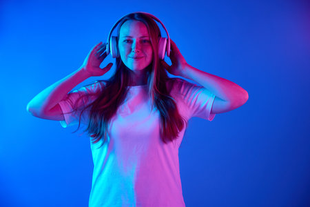 Smiling red head woman wearing headphones to listen music against gradient blue studio background in neon light. Concept of online working, hobby, shopping, freelance, delivery, technology. Adの写真素材