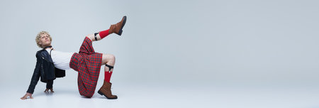 Banner. Young, handsome man in Scottish kilt and leather jacket posing against grey studio background with negative space to insert text. Concept of fashion fusion, art, uniqueness, self-expression.の写真素材