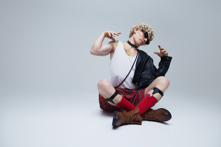Stylish young man sitting in lotus pose wearing in kilt and modern leather jacket against grey...