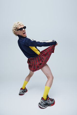 Emotional, expressive young man in plaid kilt and retro track jacket dancing, with sneakers and yellow socks against grey studio background. Concept of fashion, queer, uniqueness, self-expression. Adの写真素材