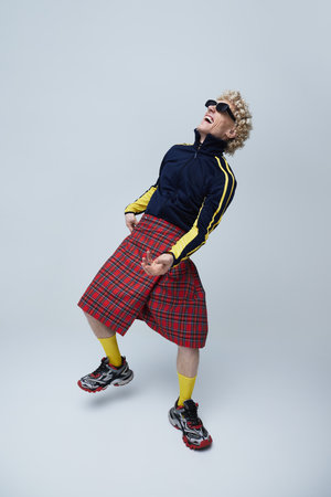 Expressive male dancer combining sportswear with a traditional Scottish skirt against grey studio...