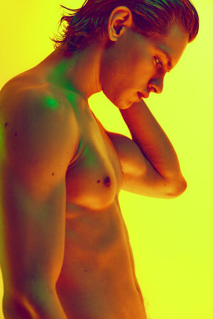 Side view. Close up photo of naked young man with strong figure in multicolored neon light against gradient yellow background. Concept of natural beauty people, fitness, male health, masculinity.の写真素材