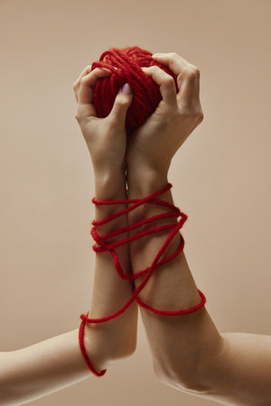 Two male and female, hands holding red ball of threads and interact with it against beige studio background demonstrating love, relationship. Concept of human touch, romantic, beauty and care.の写真素材