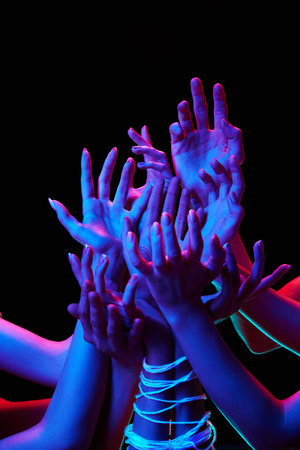 Group of hands reaching upwards in various gestures tied with strings in multicolored neon lighting...