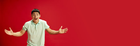 Banner. Excited young Asian man throws up his hands in delight and looking at camera against red studio background. Negative space to insert text. Concept of human emotions, fashion and beauty.の写真素材