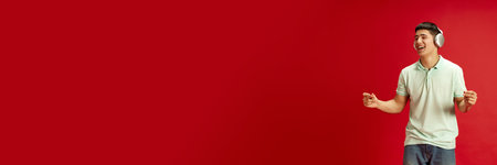 Banner. Young Asian man dancing while listening music in headphones against red studio background...