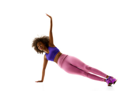 Athletic African-American woman in purple activewear balancing, training against white studio...