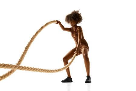 African-American woman in brown workout outfit exercising with heavy battle ropes against white...