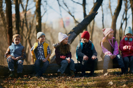 Row of kids, classmates perched on a log, clad in colorful winter wear, enjoying lesson in forest....