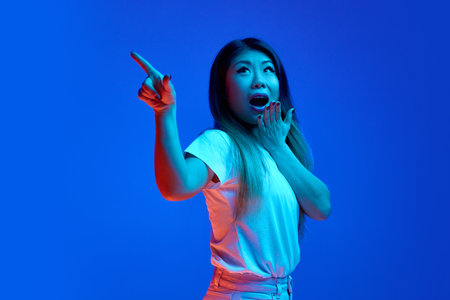 Surprised young Asian woman pointing to side and covers her mouth in surprise against blue gradient background in neon light. Concept of human emotions, self-expression, sales, shopping.の写真素材