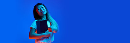 Banner. Young Asian woman, company worker shows thumb up and holding digital tablet, standing against gradient background in neon light. Concept of business, study and work, startup, sales, shopping.の写真素材