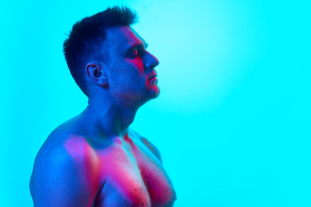 Side view portrait of handsome young man with stubble with bare shoulders looking away in neon light...