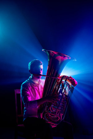 Portrait of talented musician seated, playing trumpet with soft blue-pink stage lights highlighted his silhouette. Concept of hobby and work, music festivals, concerts, symphony show, culture.の写真素材