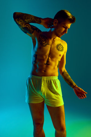 Portrait of young tattooed shirtless man posing in underwear in neon light against gradient studio background. Concept of natural beauty, fashion and style, male health and body care. Adの写真素材