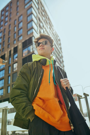 Young man in stylish sunglasses and casual attire stands confidently outside with sunlight...