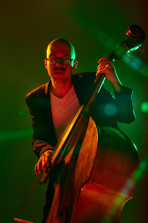 Stylish, contemporary bassist in glasses performs in red-green neon stage light against gradient...