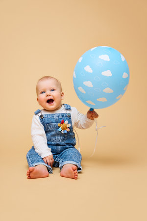 Delighted baby in denim overalls holds blue balloon adorned with clouds, and laughing against warm...