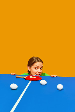 Woman peeks over table tennis table with paddle and balls, playful expression against orange background. Concept of sport, active lifestyle and healthy eating, contemporary art.の写真素材
