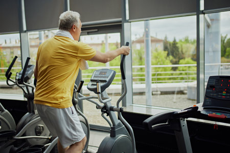 Rear view of focused senior man in vibrant sports attire with towel on shoulders work out on exercise machine at modern gym. Concept of sport, active seniors in modern life, healthy lifestyle. Adの写真素材