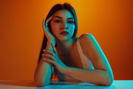 Young woman with long, dark hair elegantly posed on table, resting her head on hand in neon light against gradient background. Concept of beauty and care, cosmetology, wellness, spa treatments. Adの写真素材