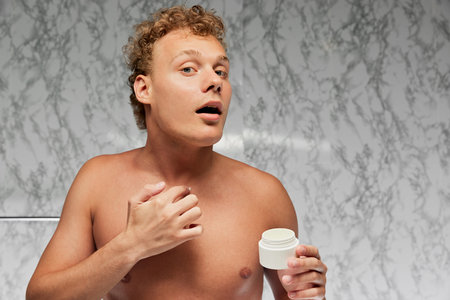 Young man with curly hair looks at camera as it mirror and applying moisturizer cream in bathroom...