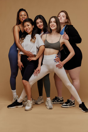 Five women of various ethnicities and body types stand together in athletic wear, smiling confidently against beige studio background. Concept of beauty and fashion, support, unity, body positivity.の写真素材