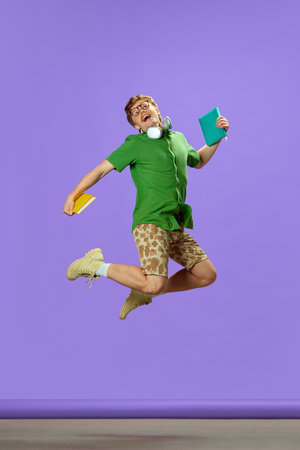 Young guy dressed casual summer attire jumping of joy and happiness holding books against purple background. Concept of education and study, skills, knowledges, graduation. Adの写真素材