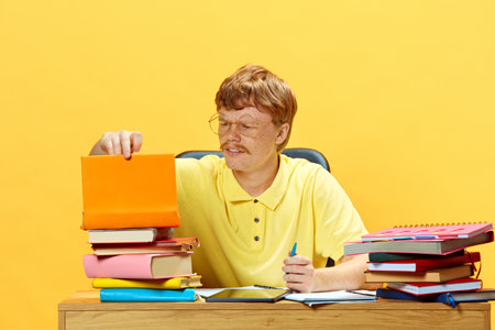 Young man sitting at desk and doing homework surrounded huge stack of books against vibrant yellow...