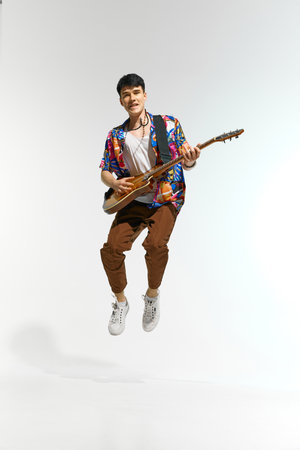 Young man in vibrant, multicolored shirt and brown pants jumps in air while passionately playing...