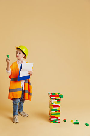 Little engineer. Child in hard hat and safety vest, with clipboard, examining tower of blocks against beige studio background. Concept of children dreams, future career, occupation, games, fantasy. Adの写真素材