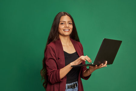 Young Indian woman holding modern digital laptop and looking at camera against green studio...
