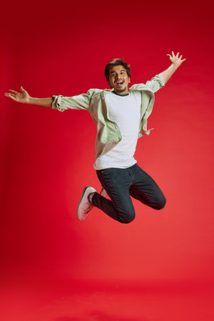 Dynamic photo of young Indian man jumping in mid-air of joy and happiness against red studio background. Positive emotions. Concept of fashion and style, human emotions, celebrating.の写真素材