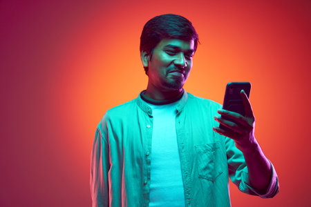 Young man looks at his phone with satisfied smile, wearing casual outfit in neon light against gradient studio background. Concept of fashion and style, human emotions, communication, shopping online.の写真素材