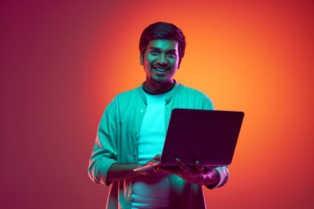Young, attractive Indian man looks at camera while using laptop in neon light against gradient...